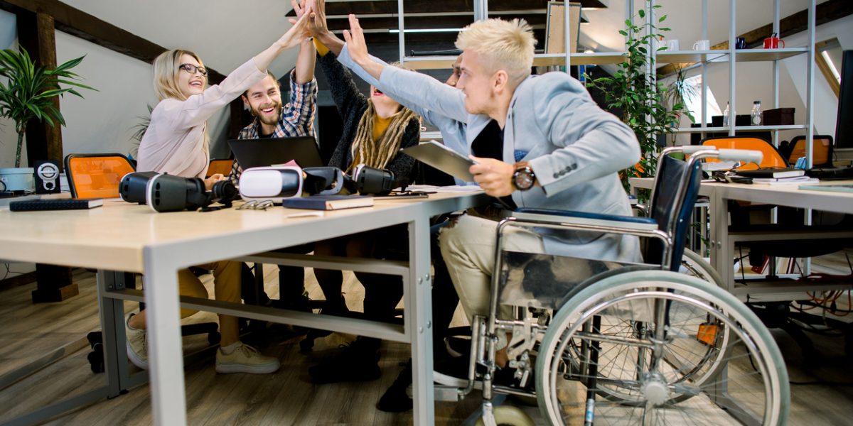 Happy multiethnic diverse young people sitting in office and giving high five each other after great collaboration, discussing strategies. Disabled male manager sitting with his colleagues at office.